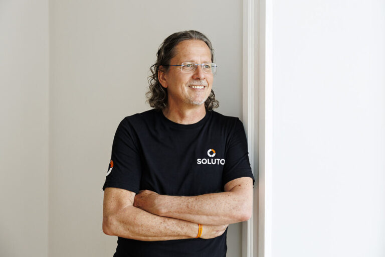 Der Gründer und Inhaber von SOLUTO lehnt entspannt und mit verschränkten Armen an einer Mauerkante, er trägt ein schwarzes Shirt mit dem Logo des Unternehmens.